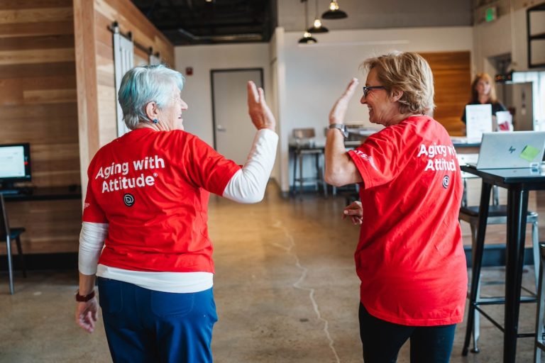 two senior women in red shirts with "Aging with Attitude" text on back giving a high five
