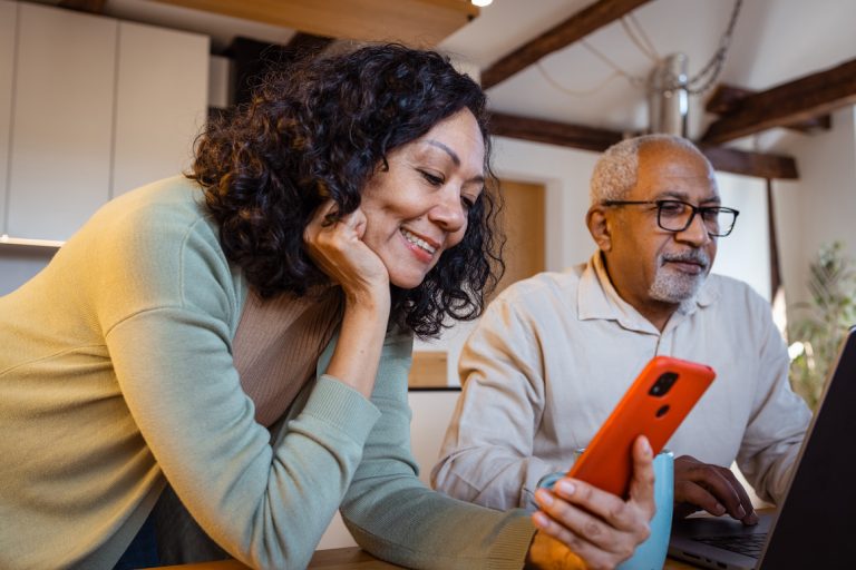 Senior couple at home. Woman is using smartphone. Man is typing on computer.