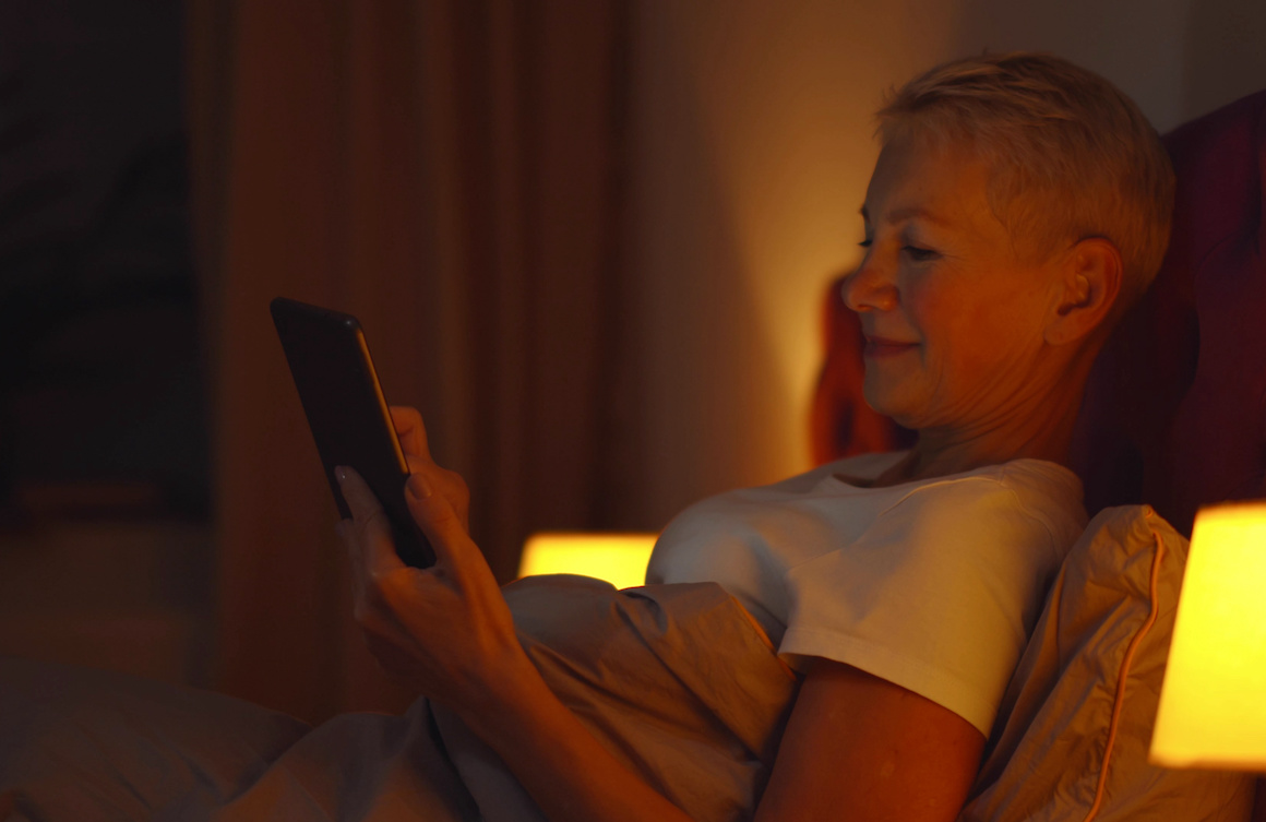senior woman using smartphone sitting on bed at home in evening