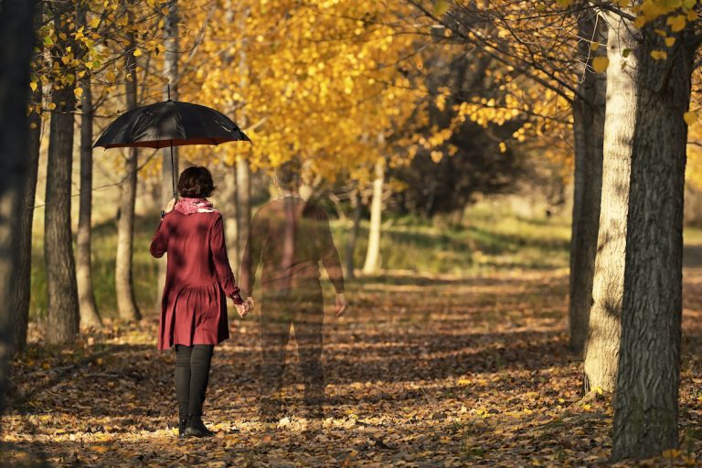 woman in red shirt holding black umbrella walking through forest in autumn while holding hand of husband. husband is translucent as if a ghost or memory.