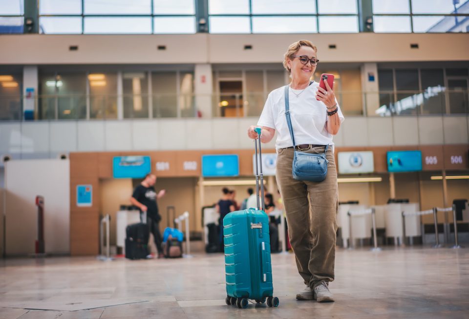 Cheerful elderly woman in glasses with a green suitcase in the lobby of the airport with a smartphone in her hands