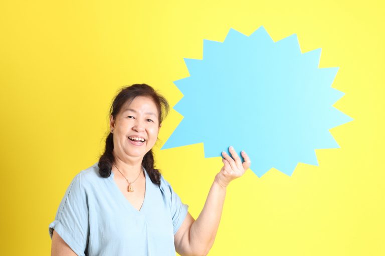 Asian senior woman wearing light blue shirt standing against yellow background holding up light blue speech bubble