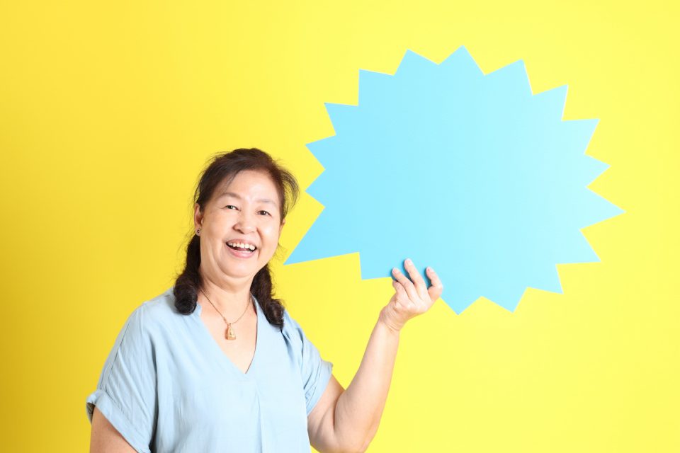 Asian senior woman wearing light blue shirt standing against yellow background holding up light blue speech bubble