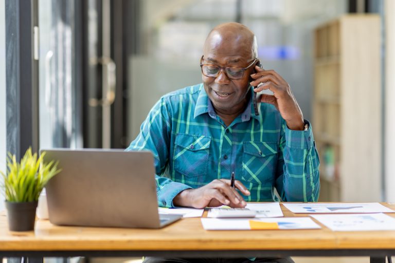 Portrait of Senior African man sitting on laptop at the home office with calculator and paperwork having a phone conversation